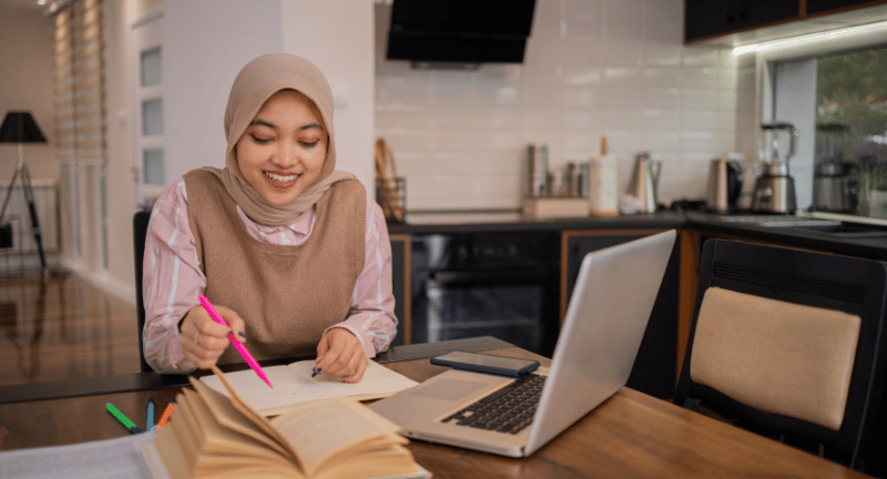 a smiling woman sits at a table with a laptop, notebook, and textbook in front of her.