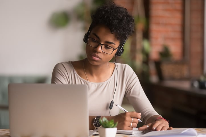 a young woman sits at a desk writing in a notepad while reading her laptop screen