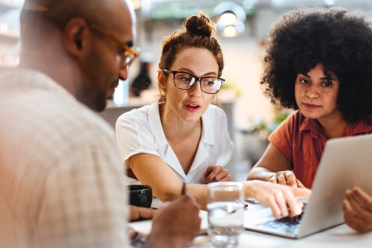 One woman points at a laptop screen with one other woman and a man next to her.