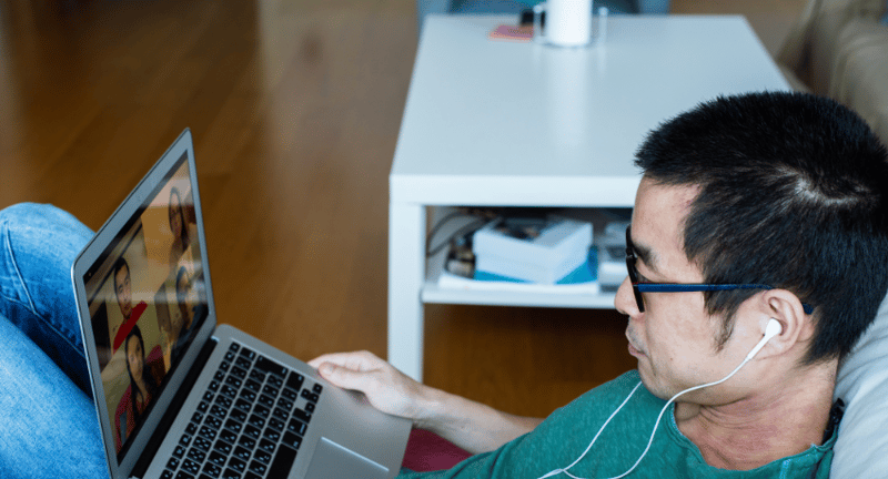 man lies on sofa chatting to other people via a video call on his laptop