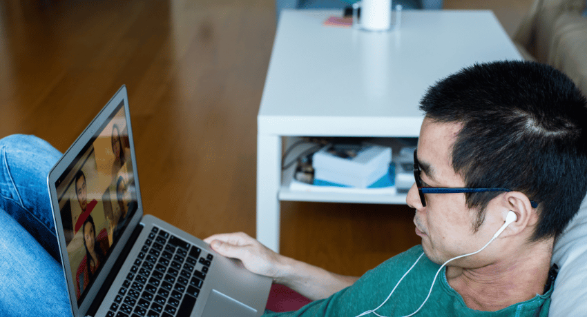 man lies on sofa chatting to other people via a video call on his laptop