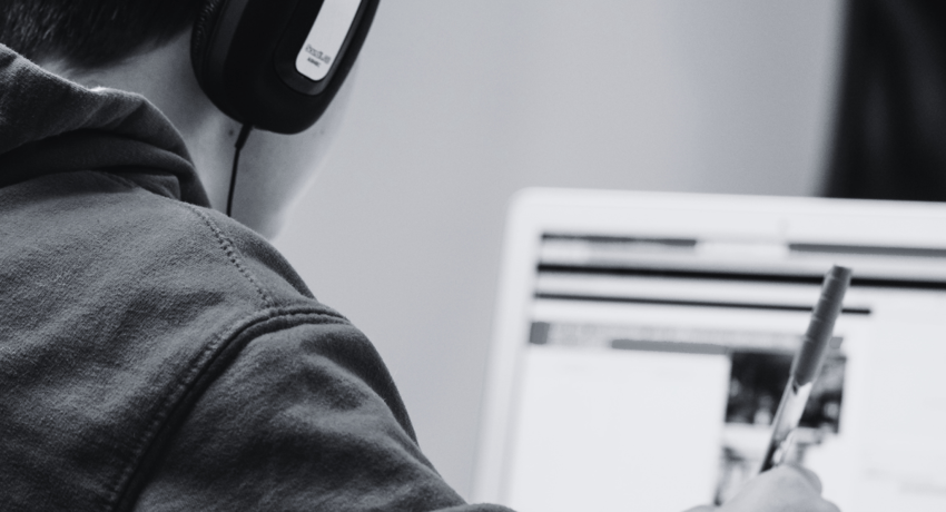 young person wearing headphones while on laptop and taking hand-written notes