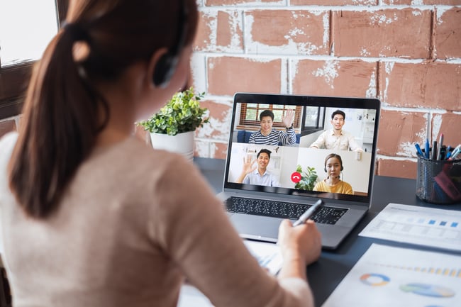 young woman sits at desk facing laptop while on a video call. There are 4 other people on the call who are shown on screen.
